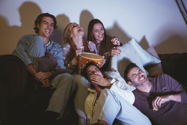 cinco pessoas sentadas no sofá comendo pipoca e sorrindo olhando para a televisão