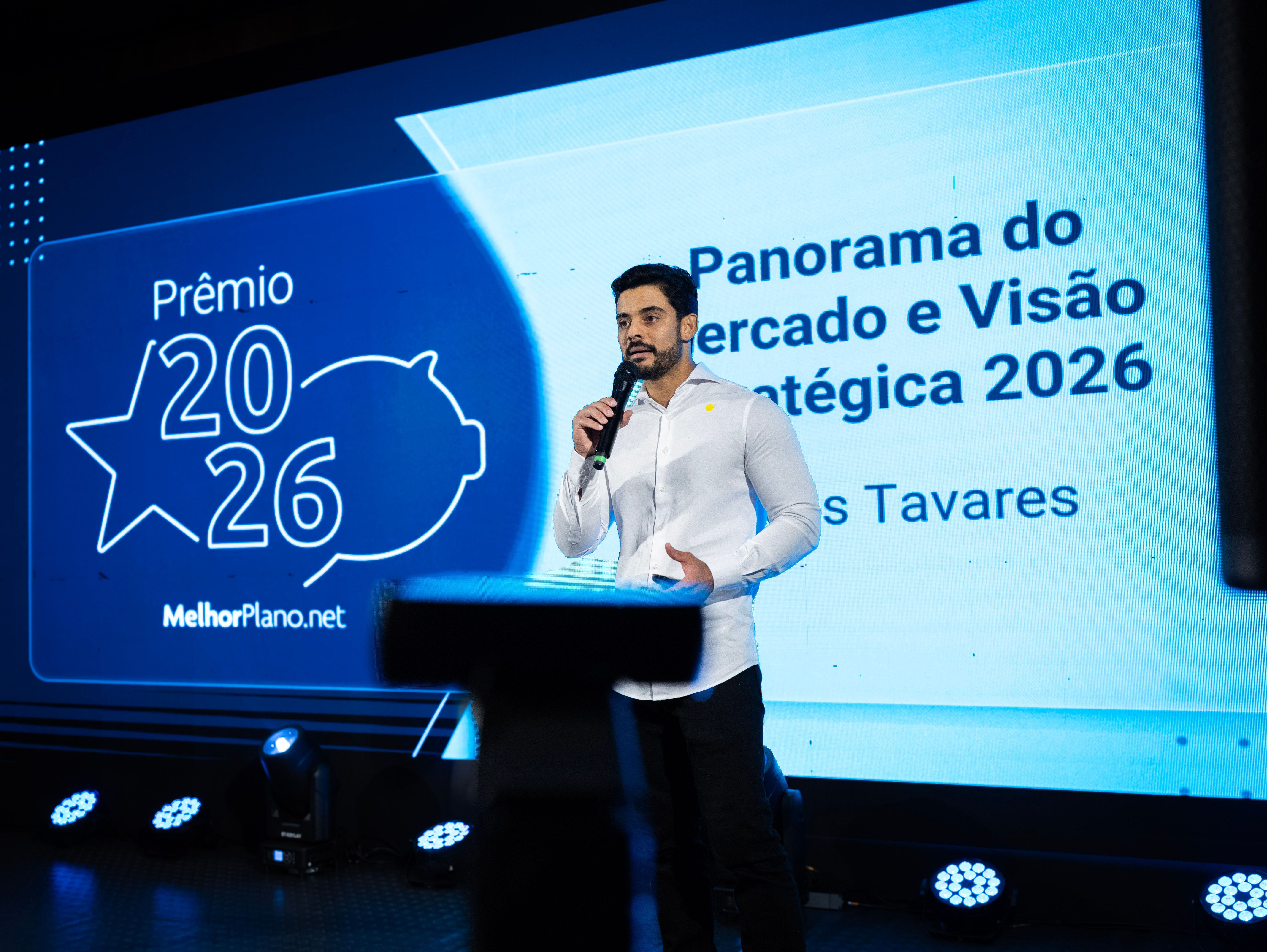 Lucas Tavares veste camisa social branca e calça preta, e está à frente de um painel com o texto 