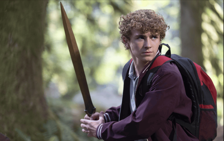 Percy Jackson em uma floresta, segurando firmemente uma espada dourada com expressão determinada. Ele veste jaqueta roxa e mochila preta com detalhes vermelhos, pronto para enfrentar um desafio.
