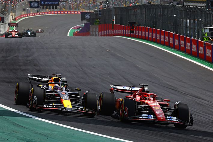 Dois carros de Fórmula 1 disputam posição em alta velocidade na pista durante uma corrida. À esquerda, um carro da Red Bull com cores azul, vermelho e amarelo; à direita, um carro da Ferrari em vermelho. A pista tem curvas amplas, proteções laterais e público ao fundo, transmitindo a emoção e intensidade da competição.